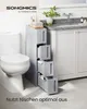 A narrow, multi-drawer storage unit is placed next to a toilet in a bathroom. The unit has several open drawers revealing toiletries. The countertop above the unit holds folded towels and decorative items. A sink and faucet are visible in the background. Produktbild-Vorschau