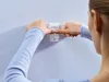 A person's hands are shown attaching a white adhesive hook to a textured light blue wall. Anteprima dell’immagine prodotto
