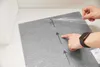 A person's hands are shown peeling protective plastic off a gray carpet tile, with arrows indicating a direction of application. Produktbild-Vorschau