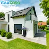 A modern white house with a balcony and solar panels on the roof, featuring a black rain barrel with purple flowers in front of a window. Produktbild-Vorschau