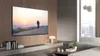 A large TV displaying a scenic image of a cyclist at sunset, mounted on a wood-paneled wall in a modern living room with a sofa and coffee table. Produktbild-Vorschau