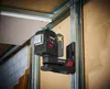 A laser level tool mounted on a metal stud wall, with green laser lines projected onto the wall and ceiling. Produktbild-Vorschau