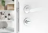 A close-up view of a white door handle and lock mechanism on a white door. The background is blurred and shows a shelf with bathroom items. Produktbild-Vorschau