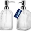 Two clear glass soap dispensers with silver pumps are shown side-by-side against a white background. A dark blue tag with the word 'orion' in light blue is attached to the right dispenser. Náhled obrázku produktu