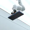 A robotic arm is holding a black rectangular object with a blue edge, possibly a screen protector, over a light-colored surface. Náhľad obrázku produktu