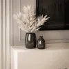 Two dark-colored vases with dried pampas grass in one, placed on a light-colored surface in front of a wall with molding. Náhled obrázku produktu