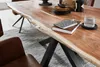 A close-up view of a live-edge acacia wood dining table with black spider legs, set in a dining room with a textured rug and a leather chair. Produktbild-Vorschau