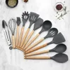A set of 12 silicone kitchen utensils with wooden handles arranged in a fan shape on a marble-like surface. Small bowls of herbs and spices are visible in the background. Náhľad obrázku produktu