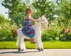 A young girl rides a plush unicorn toy outdoors in a garden setting. Anteprima dell’immagine prodotto
