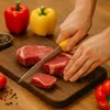 A person is cutting raw steaks on a wooden cutting board, with bell peppers and spices in the background. Produktbild-Vorschau