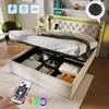 A bed with its storage compartment open, showing various items inside. A remote control is held in the foreground. Produktbild-Vorschau