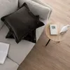 Two dark brown velvet cushion covers are placed on a light gray sofa, next to a small round wooden side table with a book and a white bird figurine. Produktbild-Vorschau
