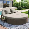 A round, rattan daybed with beige cushions and pillows is set up on a patio next to a swimming pool, surrounded by a landscaped garden. Podgląd zdjęcia produktu