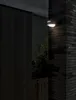 An outdoor wall light is mounted on a brick pillar next to a dark wooden fence. Produktbild-Vorschau