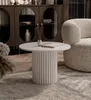 A round coffee table with a fluted base is placed on a rug in a living room setting, next to a textured armchair. Voir les images du produit