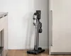 A cordless vacuum cleaner is standing upright on a wooden floor next to a white wall and a cabinet. Anteprima dell’immagine prodotto