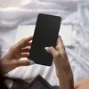 A person's hands holding a black smartphone in a bedroom setting. Náhľad obrázku produktu