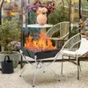 A portable grill with flames is set up in a greenhouse or sunroom with chairs and plants. Produktbild-Vorschau