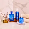 Three blue bottles of hair product and a diffuser are arranged on a marble countertop with towels in the background. Anteprima dell’immagine prodotto