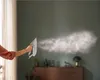 A person is holding a steam iron, releasing a cloud of steam into a room with a dresser and a lamp in the background. Voir les images du produit