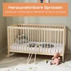 A wooden baby crib with bedding and a stuffed animal on the floor. Produktbild-Vorschau