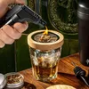 A hand is using a torch to light wood chips in a smoking apparatus placed on top of a glass of whiskey. Voir les images du produit