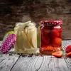 Two jars of preserved fruit, one with pears and the other with strawberries, sit on a wooden surface with scattered cloves and strawberries. Produktbild-Vorschau