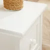 A close-up view of a white bedside table with a woven basket on top and a drawer handle visible. Produktbild-Vorschau