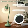 A green table lamp with a flower-shaped lampshade, displayed on a wooden table with some books and plants in the background. The image includes callouts highlighting product features. Produktbild-Vorschau