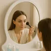 A woman applying makeup in front of a mirror, with toiletries on a shelf below. Voir les images du produit