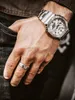 A close-up shot of a person's hand wearing a silver watch and ring, with their hand in their pocket. Voir les images du produit