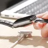 A hand is holding an Apple Watch with a black braided band, about to place it on a magnetic charging puck. A laptop is visible in the background. Produktbild-Vorschau