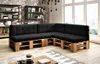 A sectional sofa made from wooden pallets with black cushions, set up in a living room with shelves and a large window. Produktbild-Vorschau
