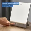 A hand is shown pressing a button on a white light therapy lamp, which is placed on a wooden table. Podgląd zdjęcia produktu