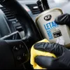 A gloved hand is cleaning a car's leather steering wheel with a yellow microfiber cloth, holding a bottle of K2 LETAN cleaner and conditioner. Podgląd zdjęcia produktu