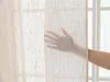 A hand is touching a sheer, beige curtain in front of a window. Produktbild-Vorschau