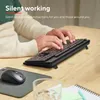 A person's hands are typing on a black wireless keyboard, with a mouse and mousepad visible on the left. The scene is set on a wooden desk. Náhled obrázku produktu