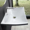 A modern white ceramic sink with a chrome faucet, set against a dark wall and a large window overlooking a natural outdoor scene with trees and fallen leaves. Produktbild-Vorschau