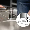 A close-up shot of a person using a socket wrench to tighten a fixture on a stainless steel sink. An inset diagram illustrates a measurement of "max. 1 mm". Produktbild-Vorschau