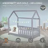 A light grey wooden cot with a house-shaped frame, decorated with a string of green pennant flags. A teddy bear is on the mattress. Icons and text describing features are at the bottom. Produktbild-Vorschau