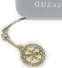 A close-up shot of a gold-colored Guess logo charm with rhinestones, attached to a phone strap, with the Guess logo visible on a grey case in the background. Podgląd zdjęcia produktu