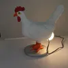 A white chicken-shaped lamp with orange feet and a red comb and wattle, standing on a gray circular base. A light bulb is visible behind the chicken, and a black cord is connected to it. Podgląd zdjęcia produktu
