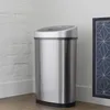 A stainless steel sensor trash can is shown in a corner next to a white brick wall and a framed picture. Produktbild-Vorschau
