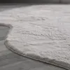A close-up, angled view of a fluffy, light gray rug with a curved edge, placed on a wooden floor. Produktbild-Vorschau