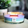 A lit candle with multiple colored layers in a glass container, placed on a wooden surface with a blurred green plant in the background. Náhled obrázku produktu