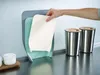 A hand is shown placing a white cutting board into a gray and mint green cutting board holder. Two stainless steel cups with wooden lids are on the right side of the counter. Náhľad obrázku produktu