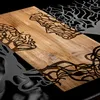 A close-up view of a wooden surface with intricate black laser-cut designs resembling tree branches and faces. Produktbild-Vorschau