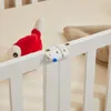 A close-up view of a white wooden crib railing with a white plastic safety latch attached. A red and white stuffed toy is visible in the background. Náhľad obrázku produktu