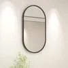 A black oval mirror with a minimalist design hangs on a white wall, illuminated by soft spotlights. A small, leafy green plant is visible in the lower left corner. Podgląd zdjęcia produktu