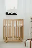 A round, wooden crib with a mattress is shown in a bright, minimalist room with sheer white curtains and decorative stars in the background. Anteprima dell’immagine prodotto